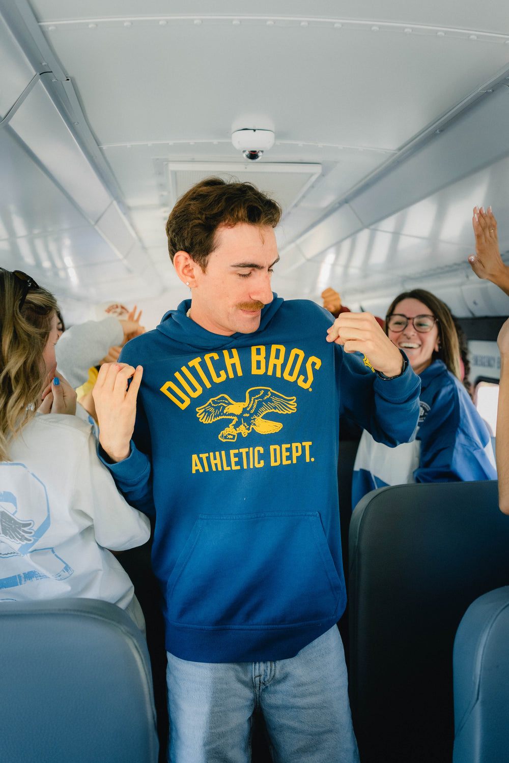 A man standing on a bus wearing a blue hoodie with yellow 'DUTCH BROS ATHLETIC DEPT.' text and a eagle graphic.