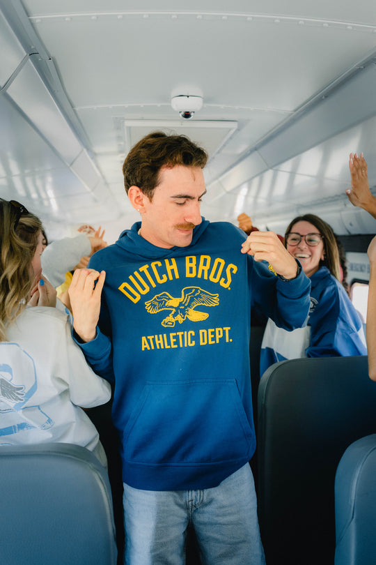 A man standing on a bus wearing a blue hoodie with yellow 'DUTCH BROS ATHLETIC DEPT.' text and a eagle graphic.
