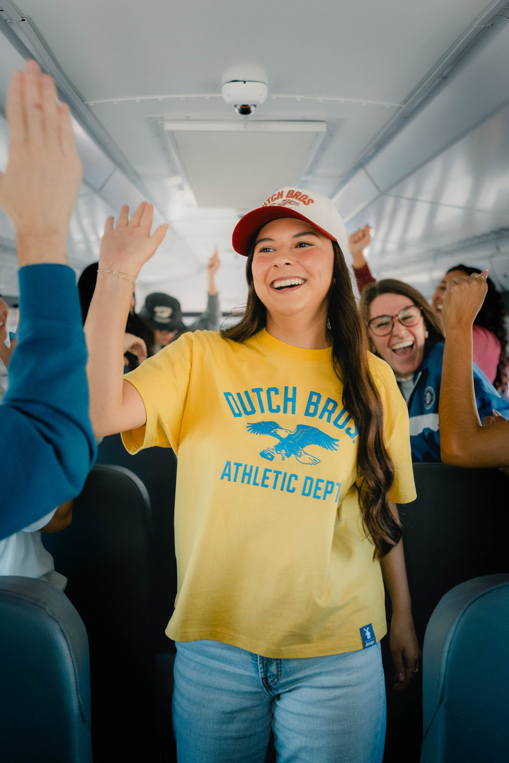 A woman standing in a bus aisle wearing a yellow t-shirt with blue 'Dutch Bros Athletic Dept' lettering and a red-and-white cap, high-fiving passengers.