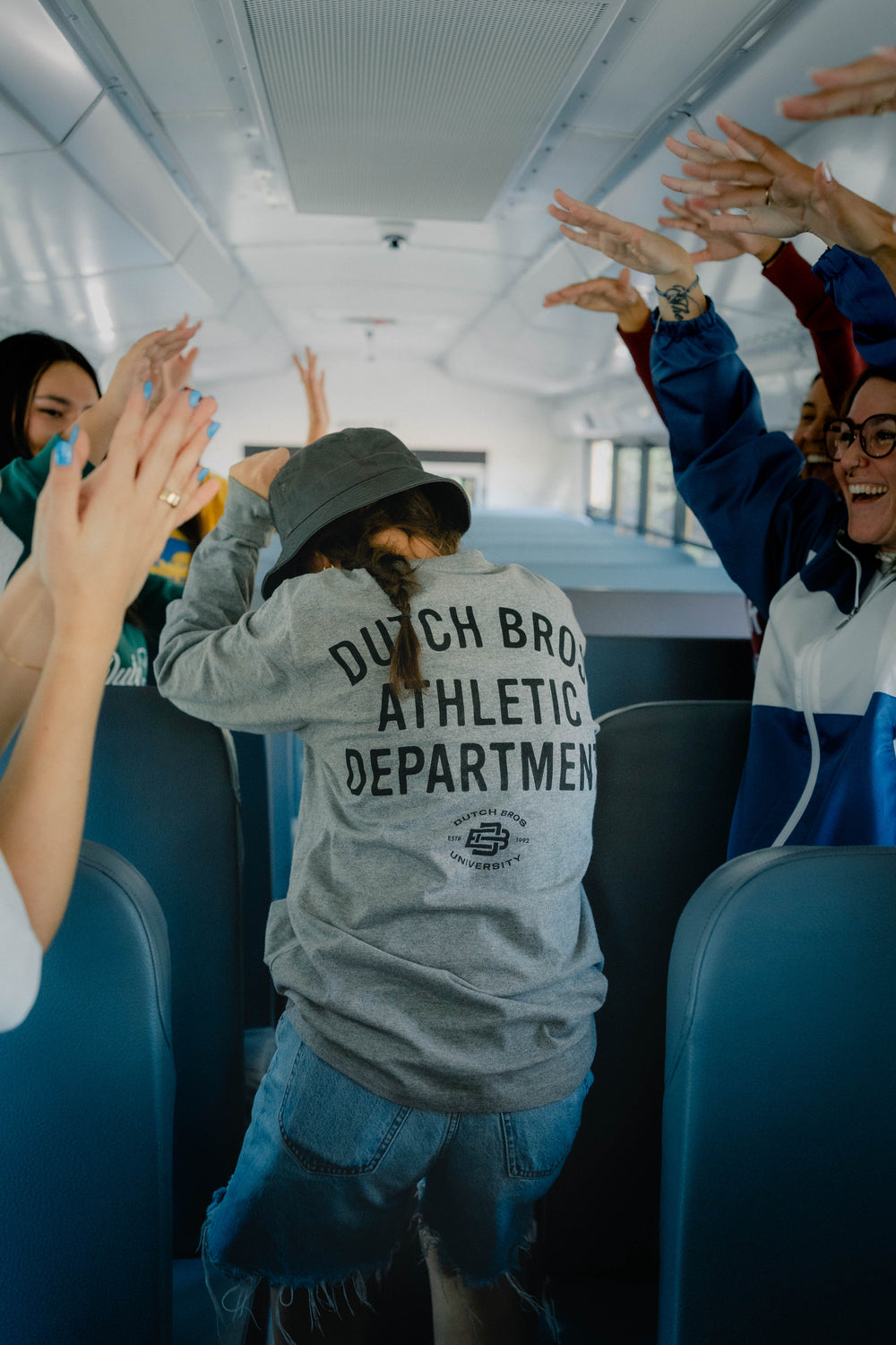 A person standing in the aisle of a bus, facing away from the camera and wearing a gray sweatshirt with 'DUTCH BROs Athletic Department' printed in black across the back. Several people around them are raising their hands in a high-five gesture.