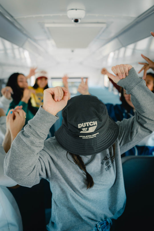 A person in a gray sweatshirt sitting on a bus or train, wearing a black bucket hat with 'DUTCH 92' printed on the front. Other passengers are visible in the background, some raising their hands.