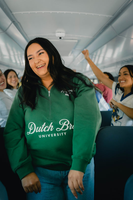 A woman smiling in a green Dutch Brue University quarter-zip sweatshirt, standing in a bus aisle with other passengers visible in the background.