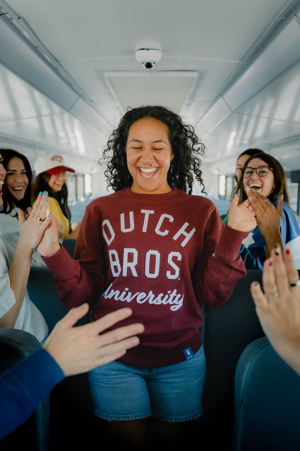 A smiling woman stands on a bus wearing a maroon sweatshirt with 'DUTCH BROS University' printed in white across the front. Other people are visible around her clapping and laughing inside the bus.