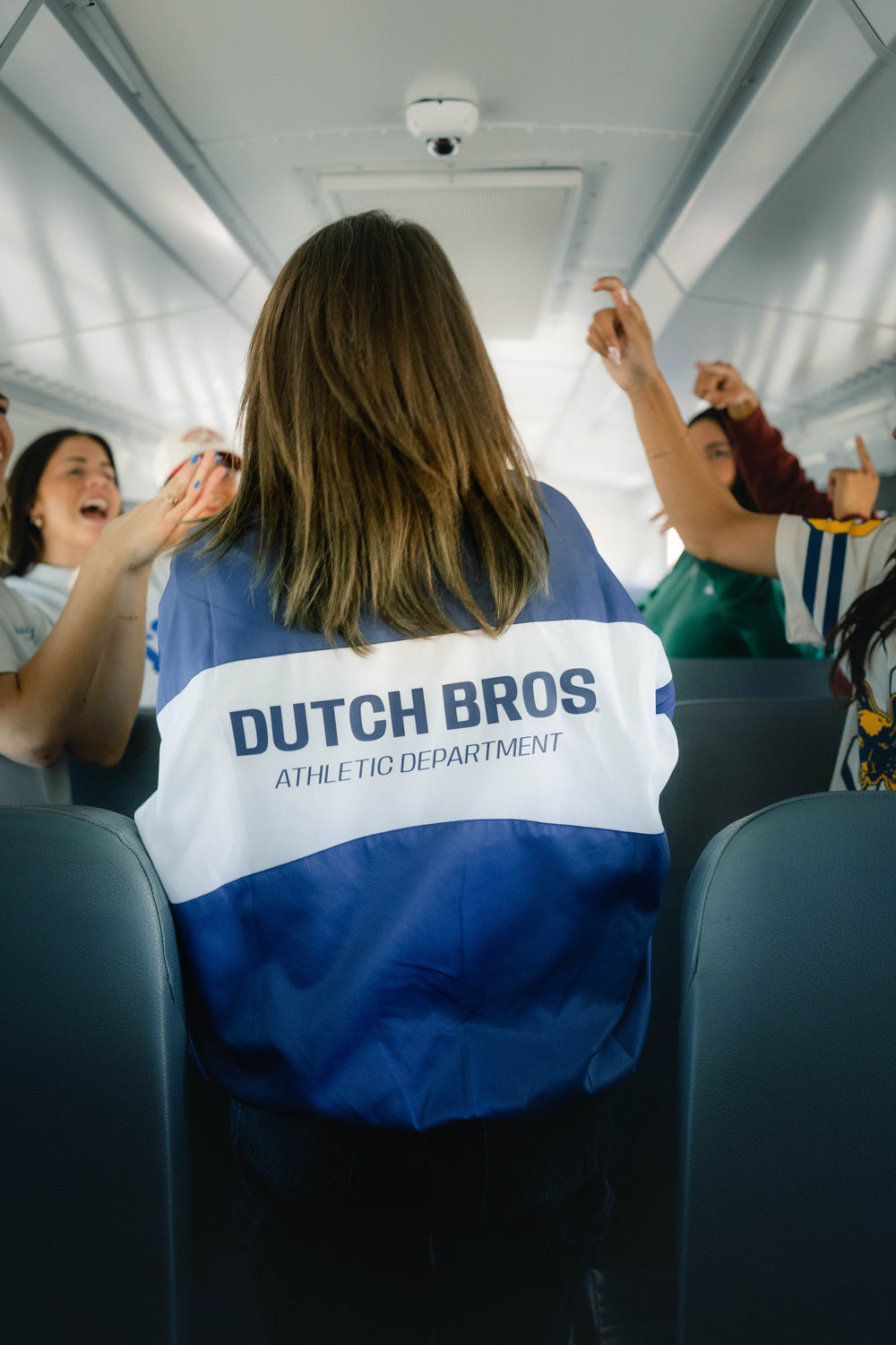 A person standing in a bus wearing a blue and white jacket with 'DUTCH BROS ATHLETIC DEPARTMENT' printed across the back. Other passengers are visible around them, some clapping and cheering.