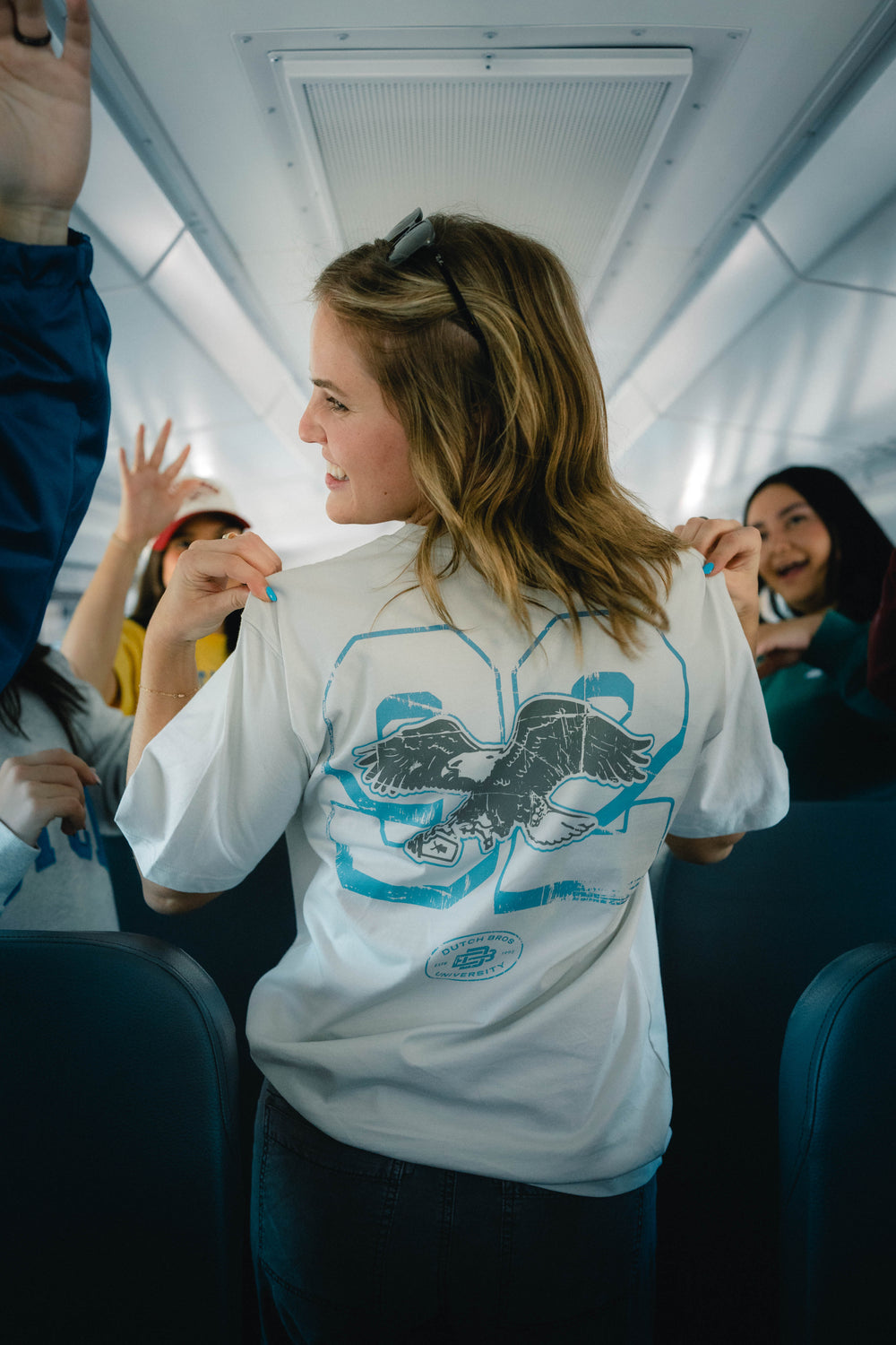 A woman standing on a bus wearing a graphic t-shirt with a blue eagle design on the back, looking over her shoulder in the bus with other passengers visible. 
