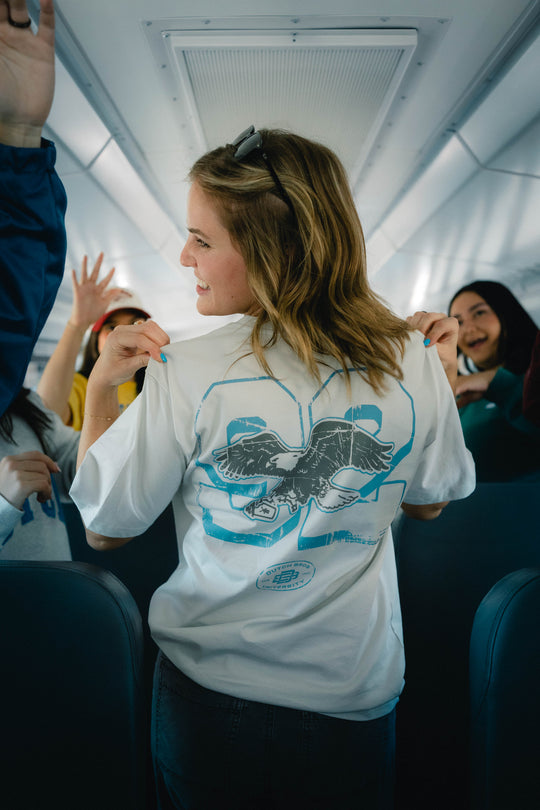 A woman standing on a bus wearing a graphic t-shirt with a blue eagle design on the back, looking over her shoulder in the bus with other passengers visible. 