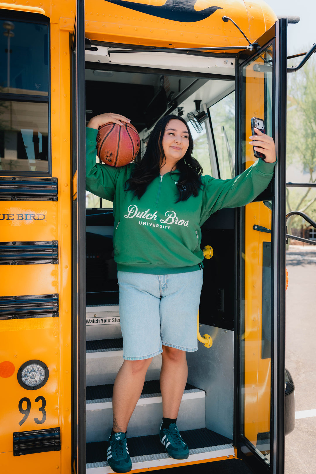 woman wearing half zip dutch bros university sweater with a basketball taking a selfie on the bus.