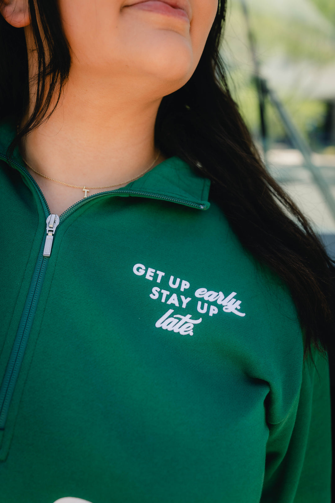 A close-up of a person wearing a dark green quarter-zip pullover with white embroidered text reading 'GET UP early STAY UP late.' on the chest.