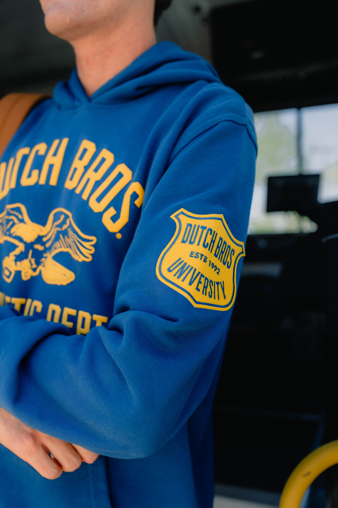 A close-up of a person wearing a royal blue hoodie with yellow 'Dutch Bros' lettering and a yellow shield patch on the sleeve, photographed inside a vehicle.