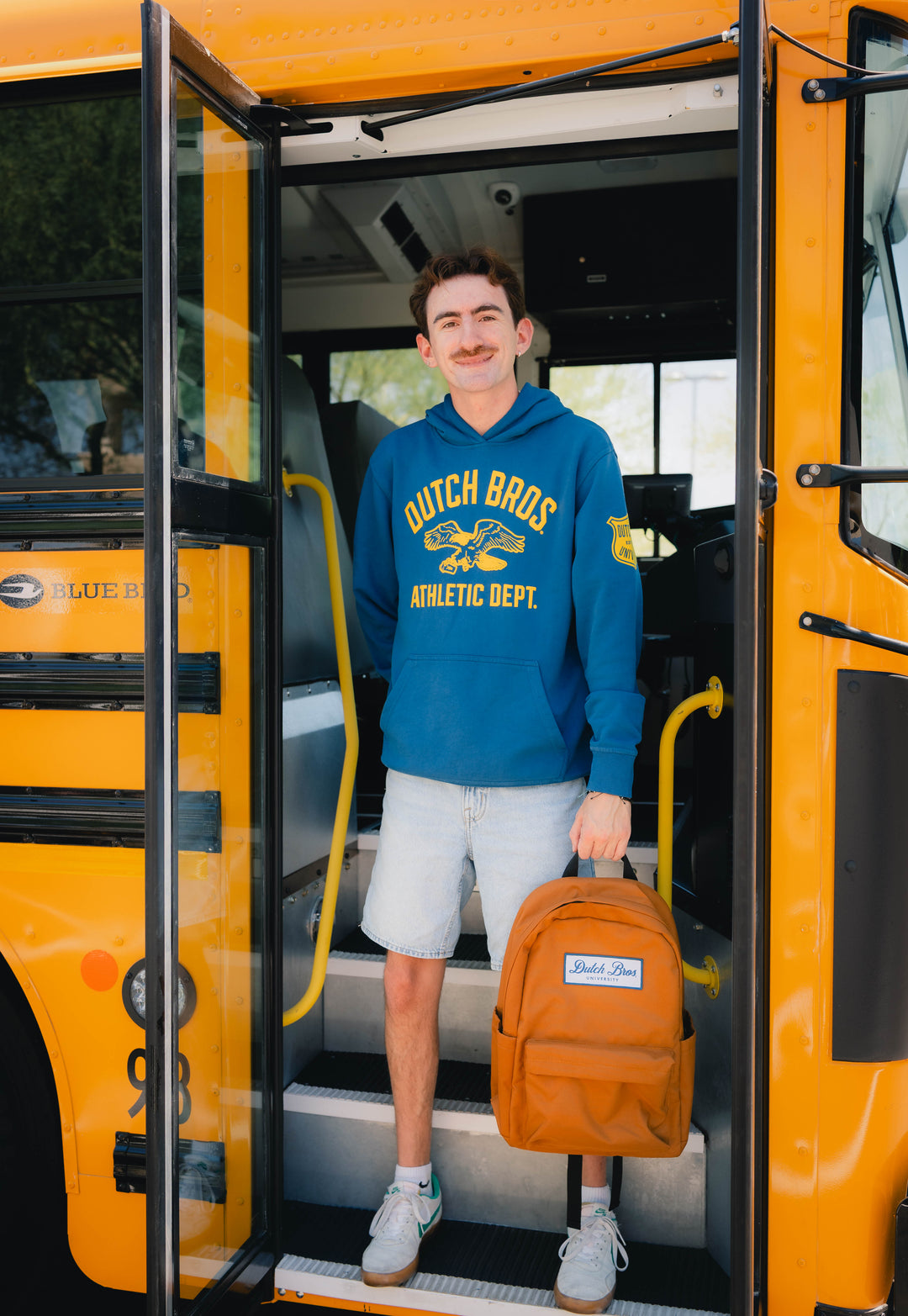 A young man stands in the doorway of a yellow school bus, holding an orange backpack. He is wearing a blue hoodie with 'Dutch Bros.