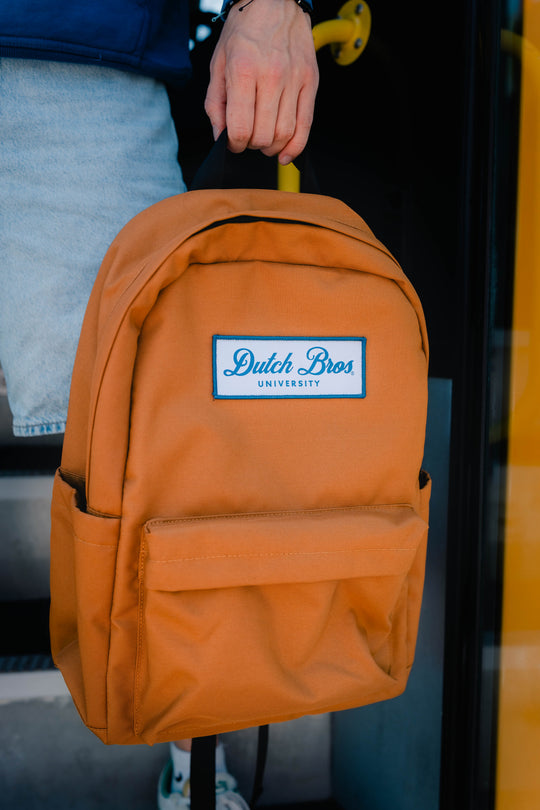 An orange canvas backpack with a white and blue 'Dutch Bros University' logo patch on the front, held by a person wearing light blue jeans.