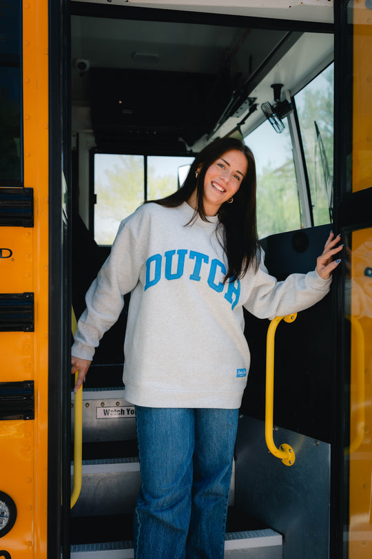 A woman stands in the open doorway of a yellow school bus, wearing a light gray sweatshirt with 'DUTCH' printed in blue across the chest and blue jeans.