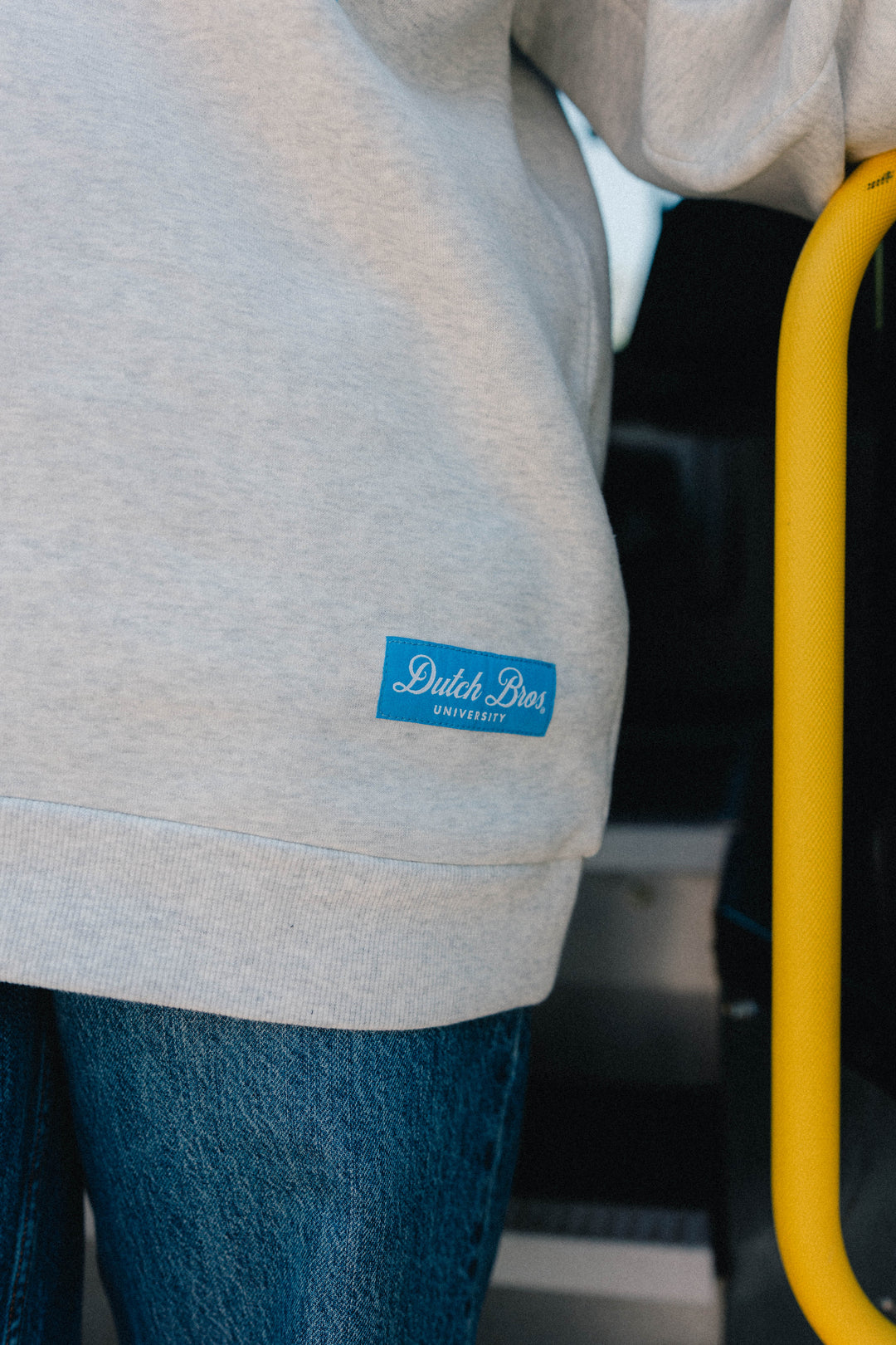 Close-up of a person wearing a light heather gray sweatshirt with a blue 'Dutch Bros University' patch on the lower left side, paired with blue jeans and standing next to a yellow bus grab handle.