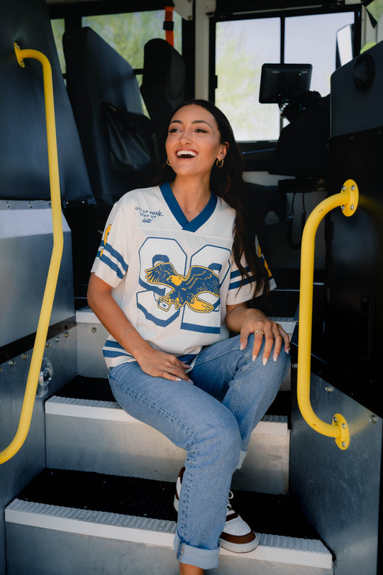 A woman sitting on the steps inside a school bus, wearing a white V-neck athletic jersey with blue and yellow details, blue jeans, and white sneakers.