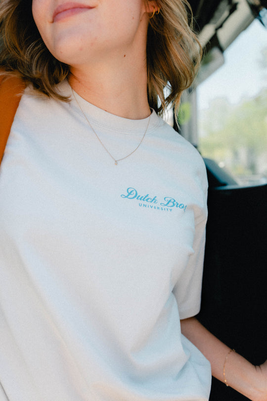 A close-up lifestyle shot of a person wearing a gray crewneck t-shirt with 'Dutch Bros UNIVERSITY' printed in light blue on the left chest, set against a blurred background of a vehicle interior.
