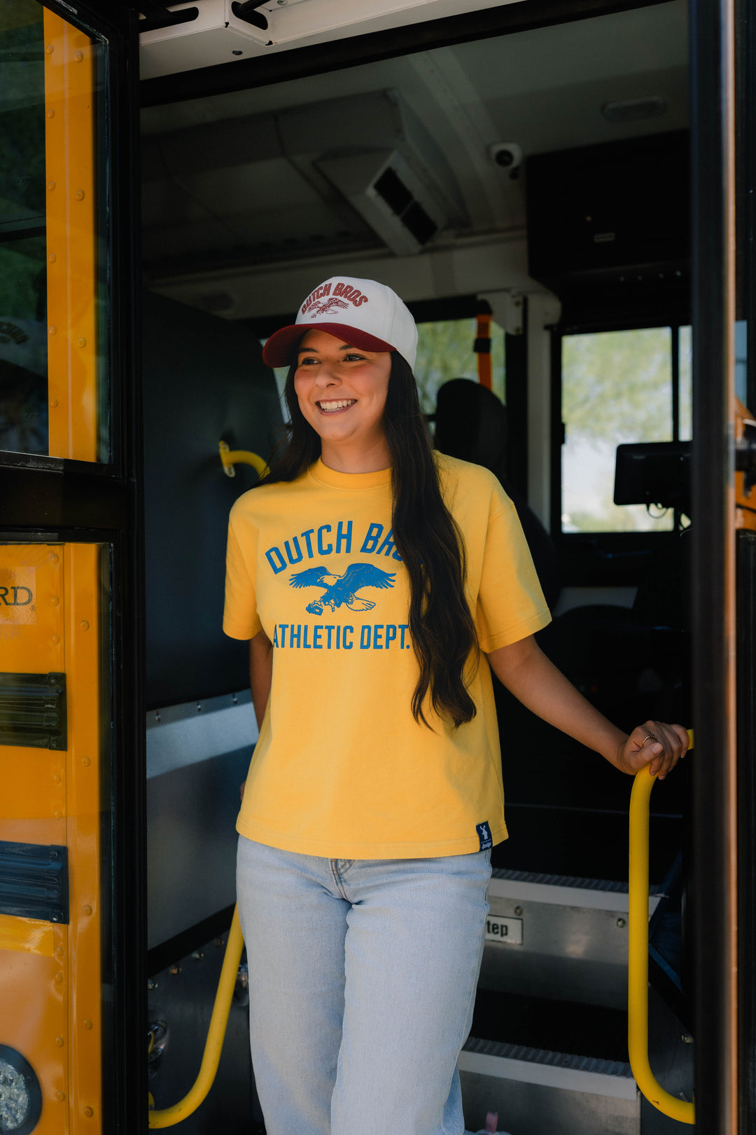 A woman smiling and stepping out of a yellow school bus wearing a yellow t-shirt with 'DUTCH BROS ATHLETIC DEPT' and a blue eagle logo, paired with light blue jeans and a white baseball cap.