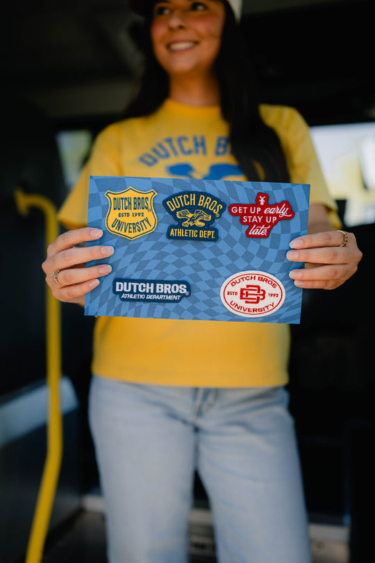 A woman in a yellow 'Dutch Bros.' t-shirt and light blue jeans holds a blue printed card featuring multiple Dutch Bros patches with different graphics on them.