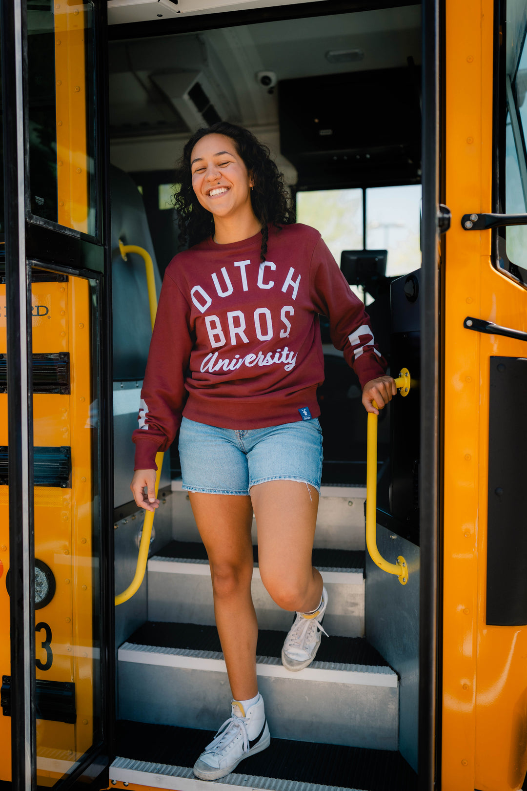 A woman smiling as she steps down the stairs of a yellow school bus, wearing a maroon sweatshirt with 'DUTCH BROS University' printed in white on the front, paired with denim shorts and white sneakers.
