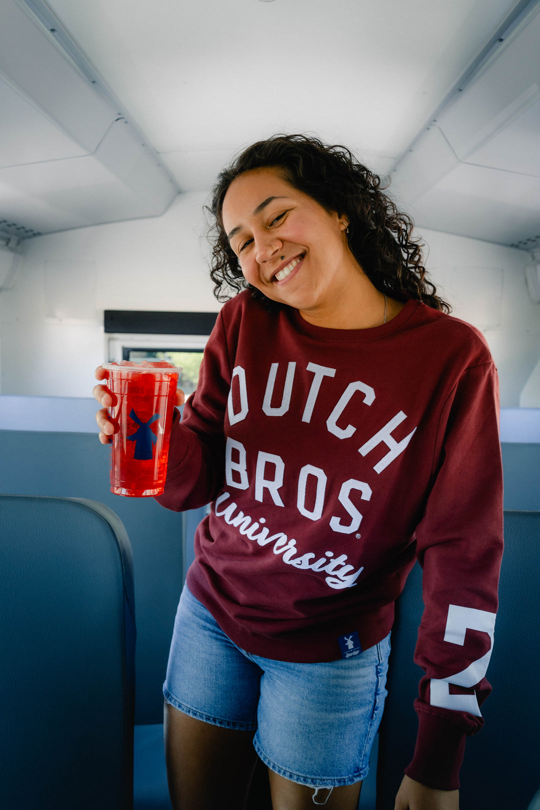 A smiling woman with curly dark hair stands inside a vehicle or bus holding a red cup. She is wearing a burgundy crewneck sweatshirt with 'DUTCH BROS University' printed in white across the front, paired with denim shorts.