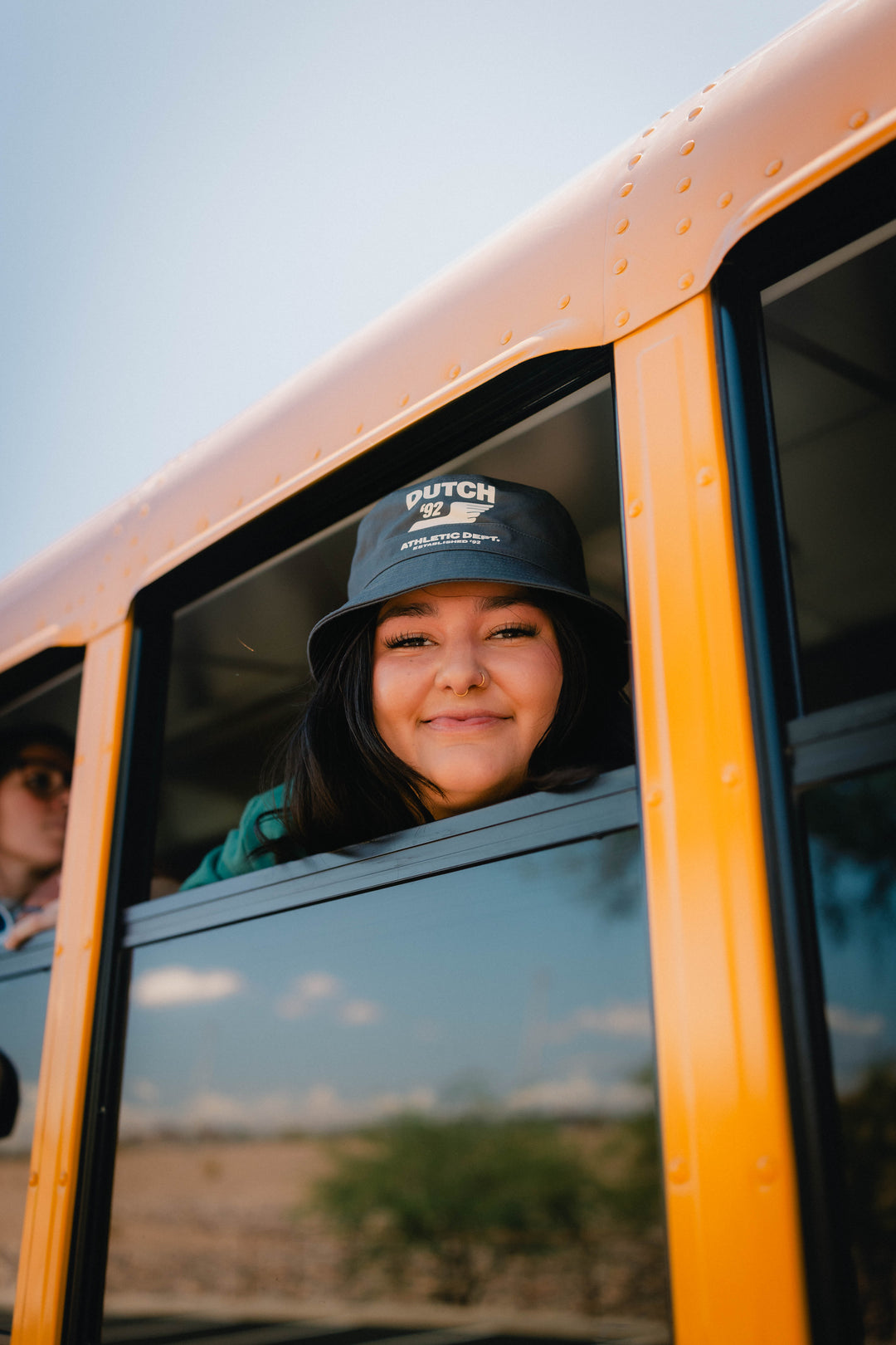 A smiling woman leans out of a yellow school bus wearing a bucket hat with the brand name 'DUTCH 92' printed in white on the front.