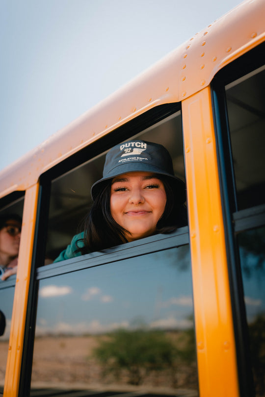 A smiling woman leans out of a yellow school bus wearing a bucket hat with the brand name 'DUTCH 92' printed in white on the front.
