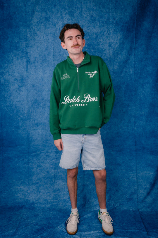 A man modeling a green half-zip pullover sweatshirt with 'Jutch Bros UNIVERSITY' printed in white on the front, paired with light denim shorts and beige sneakers, standing against a textured blue backdrop.