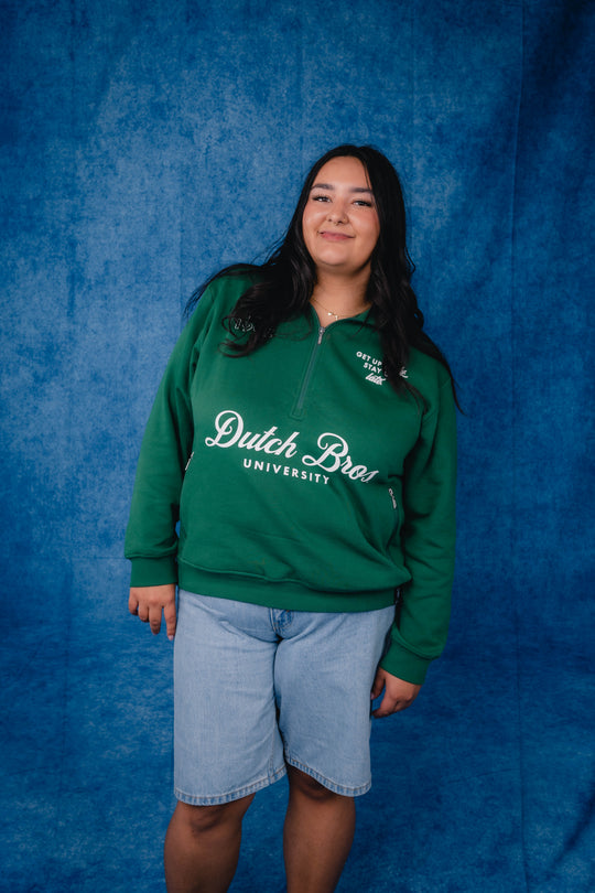 A woman wearing a green Dutch Bros. University half-zip hoodie and light blue denim shorts, standing against a textured blue backdrop.