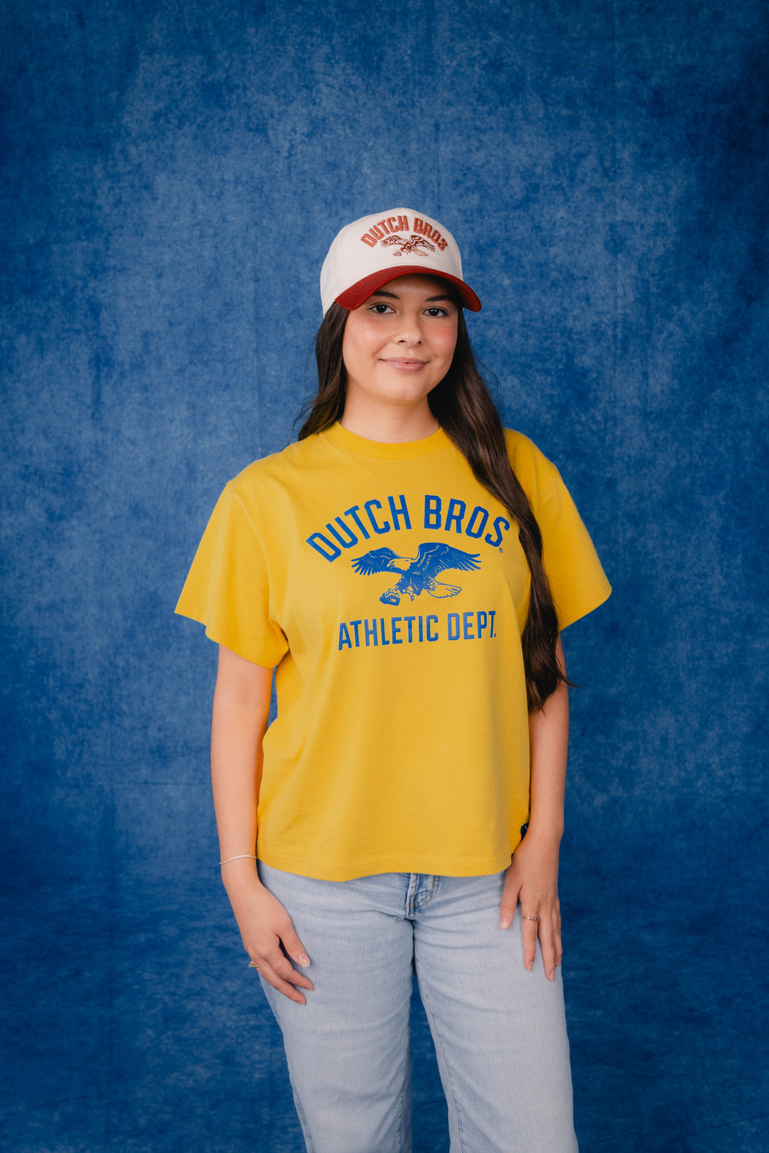 A young woman modeling a yellow short-sleeve t-shirt with a blue 'Dutch Bros Athletic Dept' eagle graphic on the front, paired with a white and red trucker hat and light wash jeans, set against a textured blue studio backdrop.