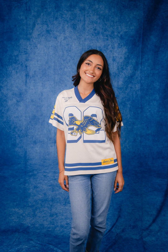 A young woman with long dark hair stands against a textured blue backdrop wearing a white and blue athletic-style jersey with a large blue number 92 and a blue-and-gold eagle graphic, paired with light blue jeans.