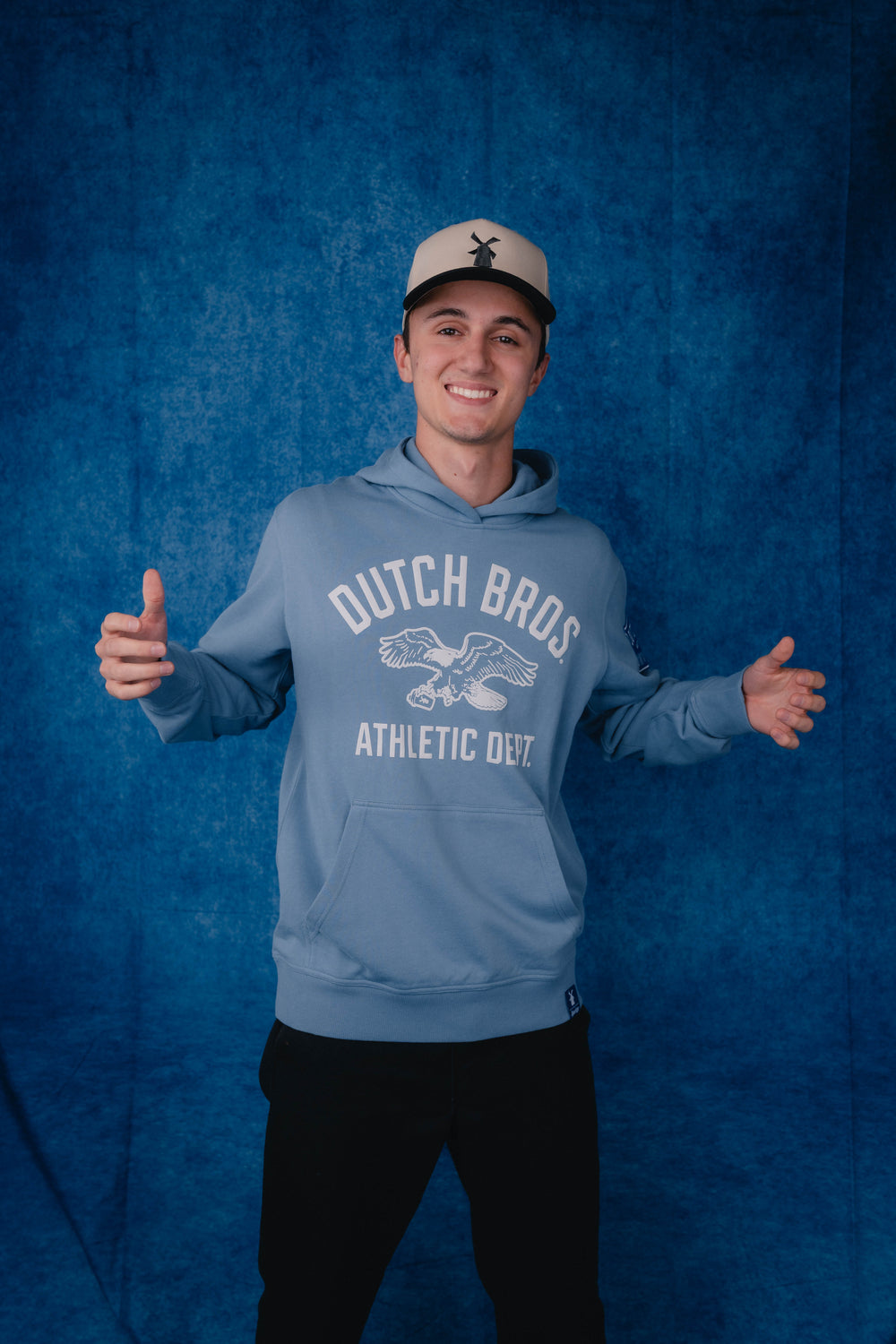 A smiling young man wearing a light blue hoodie with 'DUTCH BROS ATHLETIC DEPT' and an eagle graphic on the front, paired with a beige baseball cap and black pants, standing against a textured blue backdrop.
