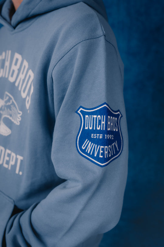 A close-up of a person wearing a light blue hoodie, showing the right sleeve with a blue and white 'Dutch Bros. University' shield patch 