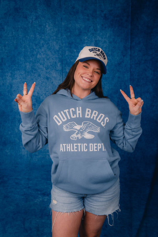 A woman posing in front of a blue textured backdrop, wearing a light blue hoodie with 'DUTCH BROS. ATHLETIC DEPT.