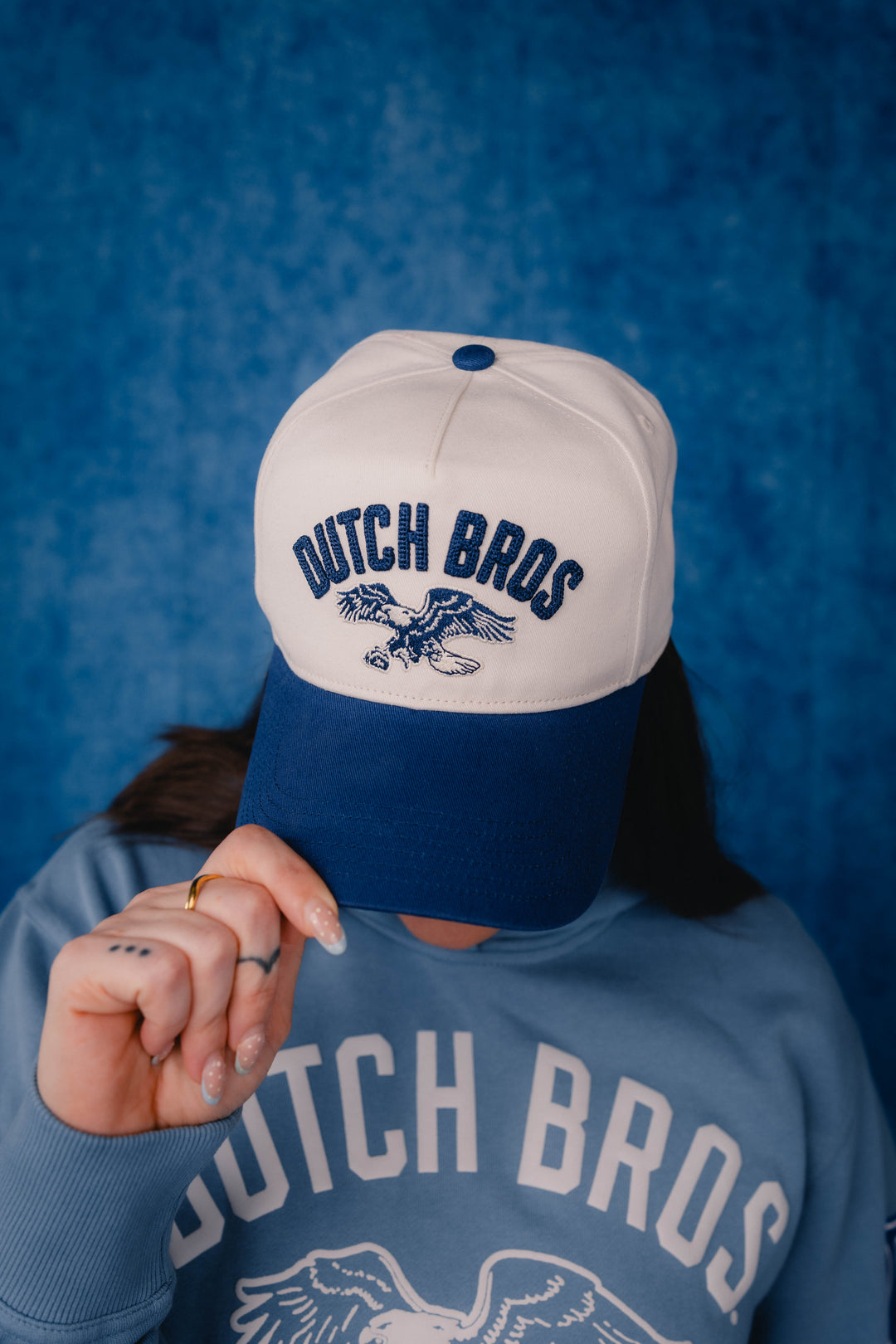 A person wearing a white and navy blue trucker hat with 'DUTCH BROS' and an eagle logo embroidered on the front, paired with a matching light blue sweatshirt against a textured blue background.
