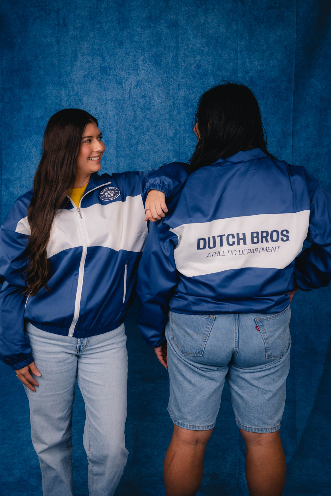 Two people wearing matching blue and white 'Dutch Bros. Athletic Department' windbreakers, standing against a textured blue backdrop.