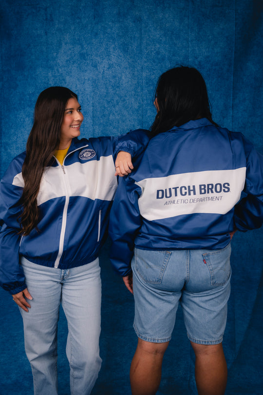 Two people wearing matching blue and white 'Dutch Bros. Athletic Department' windbreakers, standing against a textured blue backdrop.