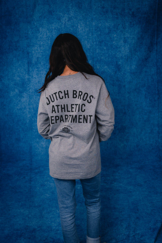 A person viewed from behind wearing a heather gray long-sleeve shirt with 'DUTCH BROS ATHLETIC DEPARTMENT' printed in black on the back, paired with blue jeans, standing against a textured blue backdrop.