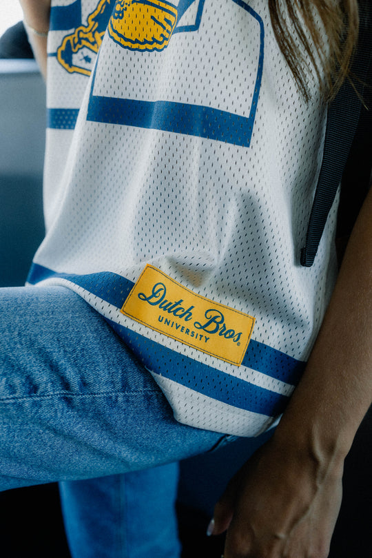 Close-up of a white mesh football jersey with blue stripes and a yellow patch reading 'Dutch Bros. University' worn by a person in blue jeans.