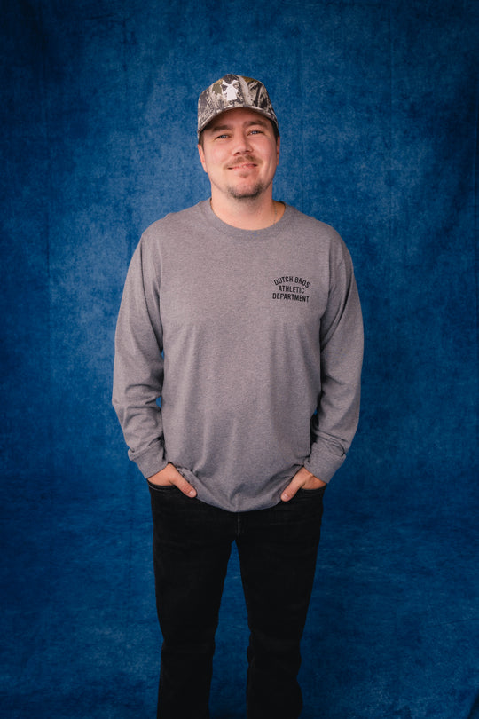 A man wearing a gray long-sleeve t-shirt with a black printed logo saying Dutch Bros Athletic Dept. on the left chest, black pants, and a camouflage baseball cap, standing against a textured blue studio backdrop.