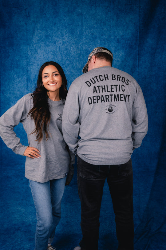 Two models wearing gray long-sleeve t-shirts against a blue textured backdrop. The woman on the left is shown from the front smiling, and the man on the right is shown from the back wearing a gray shirt with 'DUTCH BROS ATHLETIC DEPARTMENT' printed in black on the back.