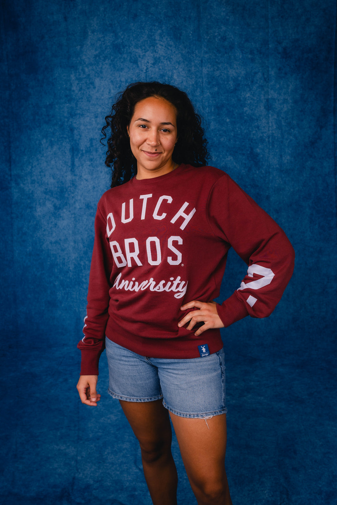 A woman modeling a maroon long-sleeve sweatshirt with 'DUTCH BROS University' printed in white across the front, paired with denim shorts, posed against a textured blue studio backdrop.