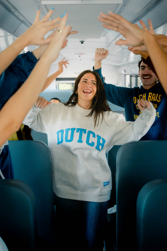 A woman in a light gray sweatshirt with 'DUTCH' printed in blue stands in a bus aisle, surrounded by classmates raising their hands. The sweatshirt features a small blue tag near the bottom right hem.