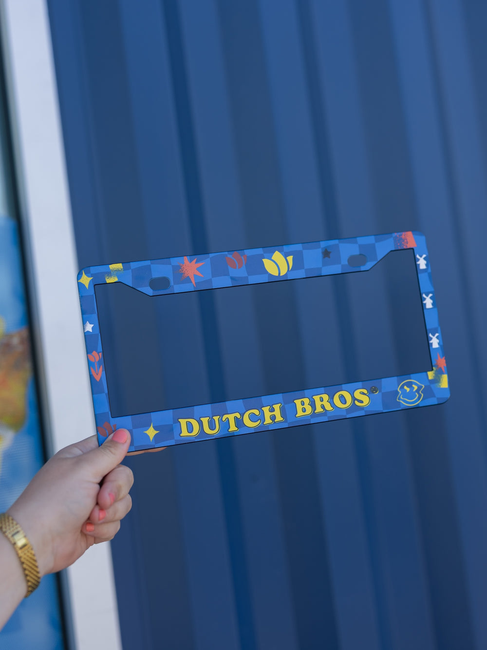 Blue license plate frame with 'Dutch Bros' text held by a hand against a blue background