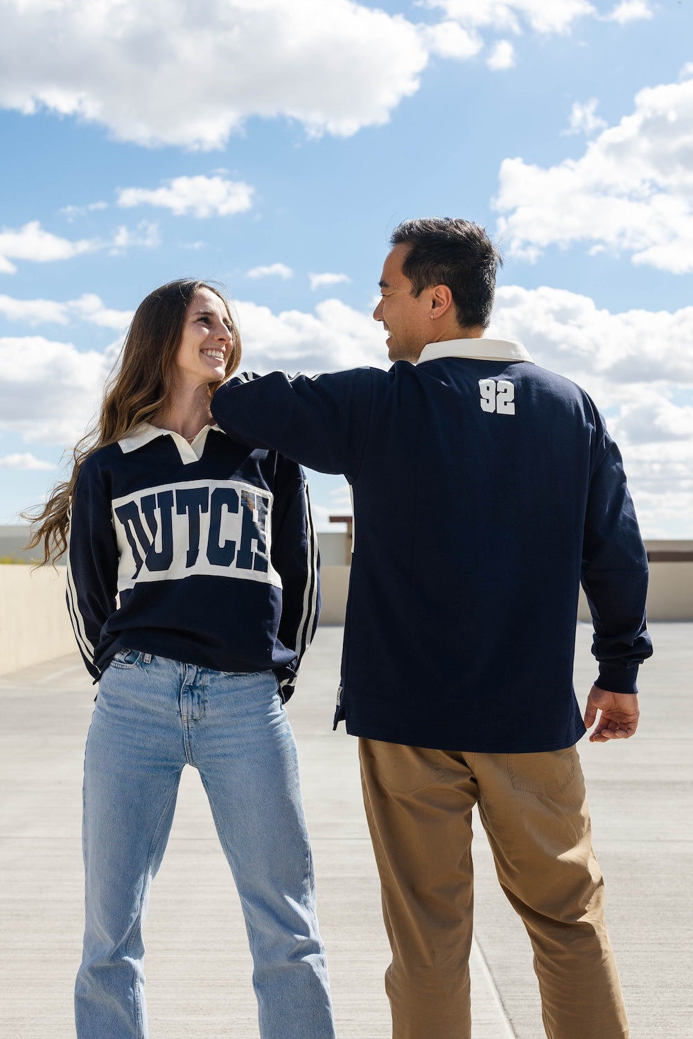 Two people wearing navy blue sweaters with 'DUTCH' printed on the back, standing outdoors under a blue sky.