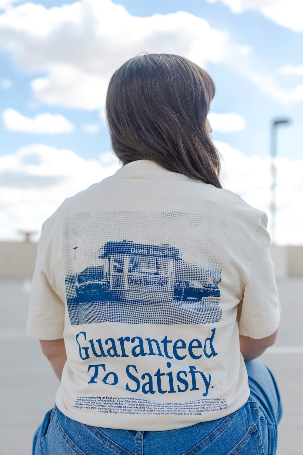 Person wearing a shirt with 'Guaranteed To Satisfy' text and graphic on the back, standing outdoors.