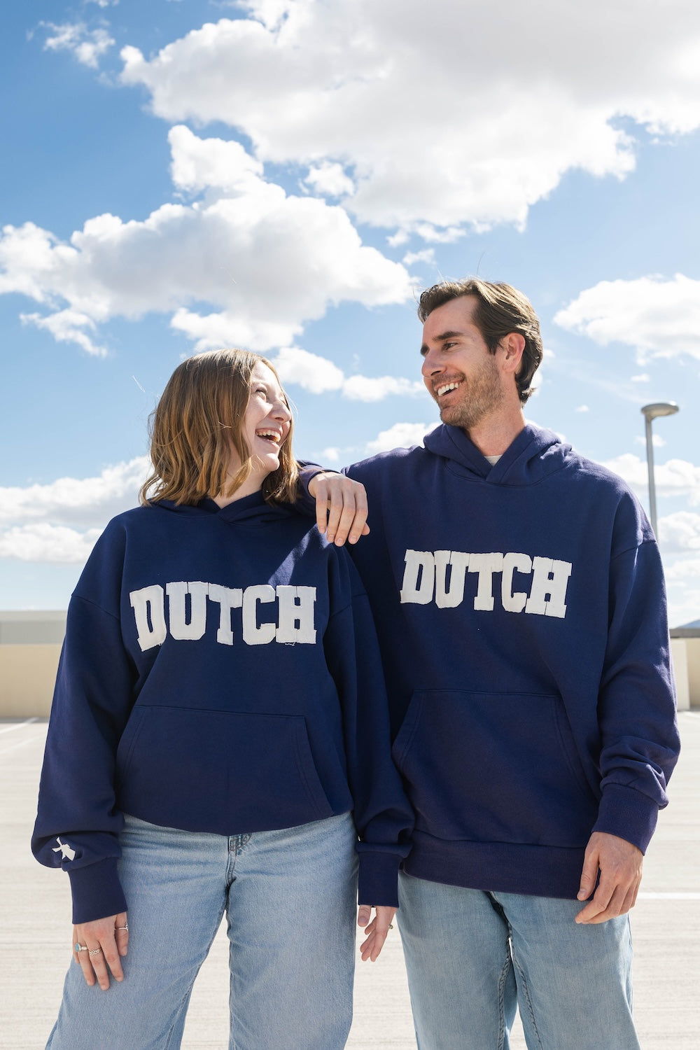 Two people wearing navy blue 'DUTCH' sweatshirts standing outdoors with a blue sky.